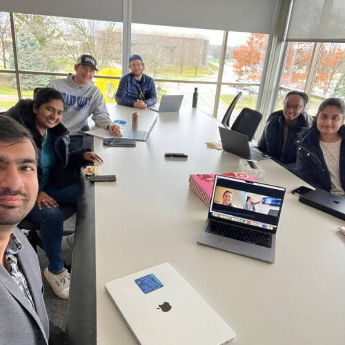 6 individuals sit along a working table smiling to a camera with laptops and phones on it.  Behind them  are large glass windows  with trees visible in the background.